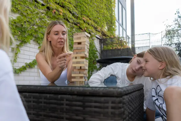 Dışarıda Jenga oynayan bir aile. Anne blok kulesini işaret ediyor, üç kızıyla yeşil, sarmaşık duvarlı evlerinin avlusunda strateji kuruyor ya da açıklıyor..