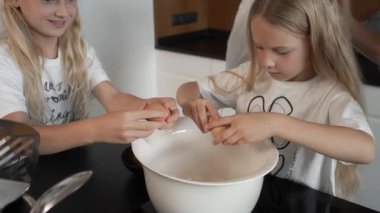 Genç kız kardeşler mutfakta işbirliği yapıyor, aile tarifi için yumurtaları dikkatlice kaseye kırıyorlar. Yaşlı bir aile üyesi onlara rehberlik ediyor. Takım çalışması ve evde temel aşçılık becerilerini öğrenme sahnesi.