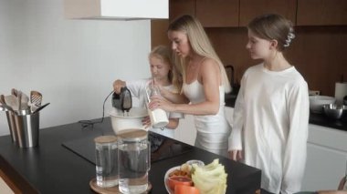 Anne ve kızları birlikte krep hamuru yapıyorlar. Annem kaseye dikkatlice un eklerken, bir kız da el mikseri kullanıyor. Kardeşler hamurun kalınlaşmasını izliyor. Mutfak ekibi çalışması ve aile eğlencesi.