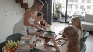 Annem kahvaltı masasında üç kızına taze pişmiş krep servis ediyor. Kızlar ev yapımı yemeklerini yiyerek meyve ve malzeme ekliyorlar. Sıcak aile sabahı atmosferi.