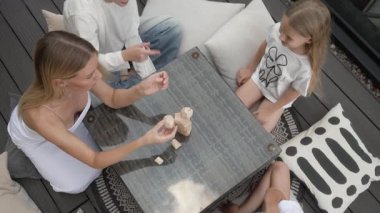 Yukarıdan aşağıya doğru: anne ve kızının elleri teras masasında eşsiz tahta bloklar ile inşa edilir. Takım çalışmasını, cesaretlendirmeyi ve yıkılmadan önce eğlenceyi paylaşarak işbirliği içinde bir kule inşa ediyorlar..