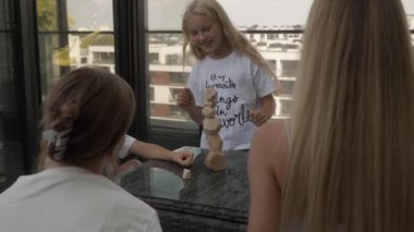 Heyecanlı kızlar şehir manzaralı terasa eşsiz ahşap blokları olan yüksek bir kule inşa etmekten gurur duyarlar. Neşeli ifadeleri bu ailenin istifleme oyununun eğlencesini ve başarısını gösteriyor..