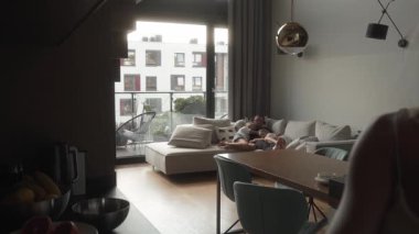 Sıcak aile sahnesi: bir baba ve iki kızı kanepede dinleniyor. Açık mutfakta, anne ailesiyle sohbet ederken yemek pişiriyor, evlerinde rahat bir ev havası yaratıyor..