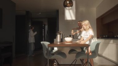 Küçük kızı mısır koçanı fırlatırken, anne buharlı tencerede yemek karıştırıyor. Büyük kızı yakınlarda dans ediyor, herkesin birbiriyle meşgul olduğu canlı bir ev ortamı yaratıyor..