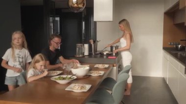 Bir aile yemeği sırasında, büyük kız babasına bir çatal uzatır, sonra da bir bardak su alır. Baba ve küçük kızı yemek yerken, anne yakınlarda yemek pişirir ve sohbet eder, sıcak, canlı bir ev ortamı yaratır..