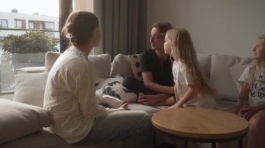 Aile baba, anne, üç küçük kız oturma odasındaki kanepede, kaliteli zaman paylaşıyorlar. Canlandırılmış bir konuşma yapıyorlar. Sıcak evlerinde jestler yapıp birlikte gülümsüyorlar..