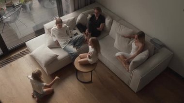 Sessiz sinema oyununda eğlenen bir ailenin yüksek açılı görüntüsü. Kızlar sırayla yerde ve masada anlamlı pozlar vererek yaratıcı ipuçlarını değerlendirir. Keyifli bir ev eğlencesi paylaşırlar..