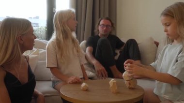 Bir aile (ebeveynler ve iki kızı) evde yaratıcı bir ahşap istifleme oyunu oynarken neşeli ve odaklanmış bir anı paylaşır. Dikkatli bir şekilde bir kule inşa ederler, dengeyi, beceriyi ve beraberliği test ederler..