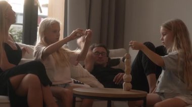 İki genç kız kardeş, ahşap denge taşları olan yüksek bir kule inşa etmeye derinden odaklanmış durumdalar. Aileleri kanepede dinlenir, sohbet eder ve kızların evdeki oyun saatlerini izlerler..