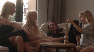 Kız kardeşlerin oyun zamanı: genç kızın tahta blok kulesi çöker. Büyük abla yardım ediyor, gülüyorlar ve kardeş takım çalışmasını ve dayanıklılığını göstererek dengeleme oyununu yeniden başlatıyorlar. Ebeveynler kanepede sohbet eder..