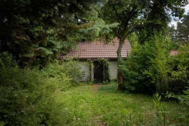 Aşırı büyümüş yeşil yapraklar ve olgun ağaçlarla kaplanmış küçük, kırsal bir kır evinin dışında. Kırmızı fayanslı bir çatısı ve asma kaplı ahşap bir kapısı vardır. Doğal, dağınık bir bahçe ortamında gözlerden uzak bir yer.