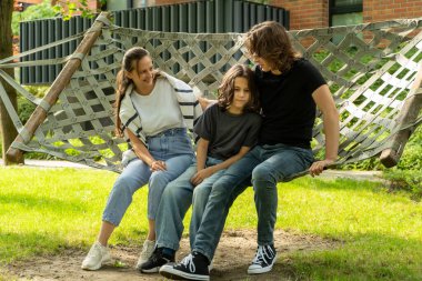 Ebeveynler oğullarıyla birlikte açık bir hamağın kenarında otururlar; baba sevgisini ve desteğini ifade ederek çocuğa sarılır. Anne gülümsüyor, bu huzurlu aile anının tadını çıkarıyor..