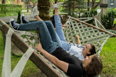 Küçük bir kardeş, büyük bir açık hava hamağında birlikte yatarken, gülen annesi ve ağabeyi ile şakalaşarak sallanıyor. Bu neşeli sahne aktif bir aile eğlencesini yakalıyor..