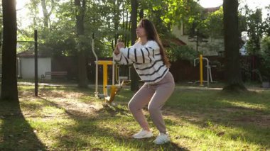 Güneşli bir park ortamında vücut ağırlığı çömelmesi yapan aktif bir kadın. Açık hava spor programının bir parçası. Bu fiziksel aktivite sağlık, bacak dayanıklılığını ve genel sağlığını teşvik eder..