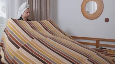 Sabah rutinine devam ederken pijama ve havlu giymiş bir kadın düzgünce yapılmış yatağın üzerine örülmüş bir yatak örtüsü serer. Düzenli ve davetkar bir yatak odası için son katmanı ekliyorum..
