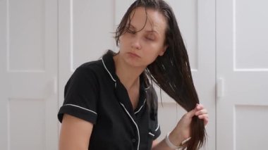 Pijamalı bir kadın uzun, ıslak saçlarını çözücü bir saç fırçasıyla dikkatlice fırçalar. Rutubetli kilitlerini titizlikle açıyor, sabah saç bakımının bir parçası olarak onları düzleştiriyor ve çözüyordu..