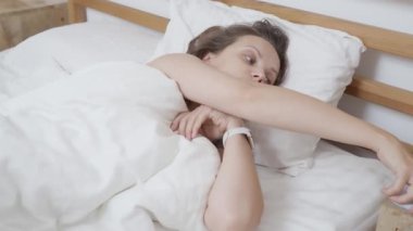 Akıllı bir bant takan kadın, yatakta uyanıyor, yastığı ayarlıyor ve akıllı telefonunu komodinden alıyor. Güne mesajları kontrol ederek ya da cep telefonuna bakarak başlıyor..