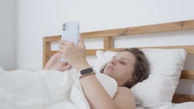 Rahat kadın akıllı telefonunu kullanarak yatakta uzanıyor, sosyal medyada geziniyor ya da haberleri okuyor. Yavaş bir sabahtan zevk alıyor, cep telefonu ve akıllı saatiyle bağlı ve bilgilendiriliyor..