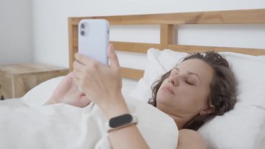Yatakta uzanan bir kadın, cep telefonunu sabah dijital programı için kullanır. Video içeriği, çevrimiçi alışveriş yapıyor olabilir ya da kalkmadan önce mesajlara cevap veriyor olabilir..
