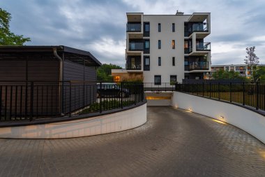 Aydınlatılmış kıvrımlı asfalt yol modern bir apartmanın otoparkına çıkıyor. Konut kompleksi içine entegre edilmiş kullanışlı, güvenli araç park olanağı sunar.