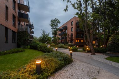 Günbatımında modern konut kompleksinin peyzajlı avlusunda aydınlık yol. Bollard ışıkları, çimenler, çalılar, ağaçlar ve apartman binaları arasında sıcak bir ortam yaratıyor. Huzurlu bir topluluk alanı.