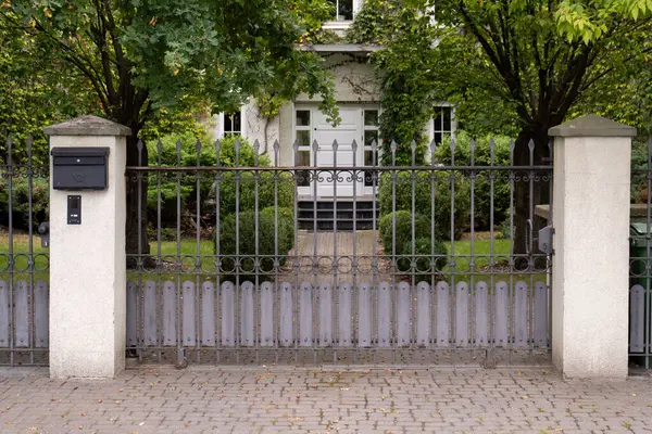 Gözlerden uzak bir eve iyi tanımlanmış bir giriş. Posta kutusunun olduğu önemli sütunlar arasında dekoratif metal bir kapı var. Manzaralı bir patika sarmaşık kaplı bir eve doğru gidiyor..