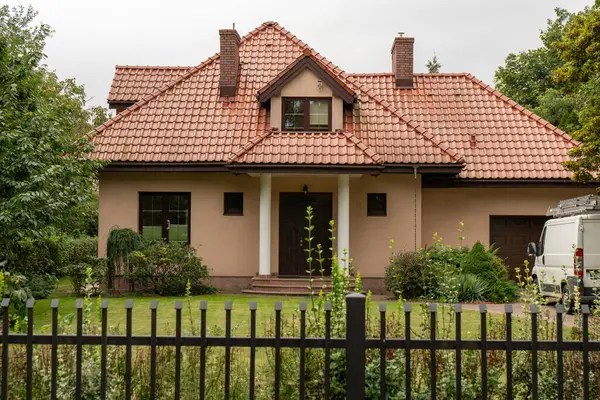 Döşeme çatılı, yatakhaneli ve giriş sütunlu geleneksel bir aile evi. Koyu metal çitler yeşil çimlerle, çalılarla çevrili. Banliyöde rahat yaşam, klasik ev cazibesi manzarası.