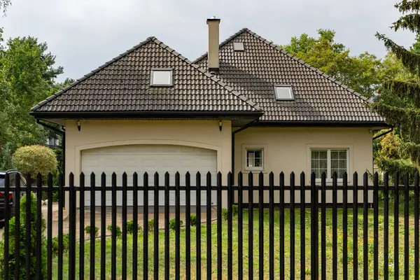 Modern aile evi, aydınlık cephe, koyu kiremitli çatı, tavan penceresi, entegre garaj. Siyah metal çitler yeşil çimlerle çevrili. Güvenli, şık bir banliyö hayatı manzarası.