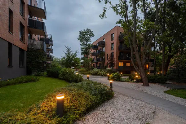 Günbatımında modern konut kompleksinin peyzajlı avlusunda aydınlık yol. Bollard ışıkları, çimenler, çalılar, ağaçlar ve apartman binaları arasında sıcak bir ortam yaratıyor. Huzurlu bir topluluk alanı.