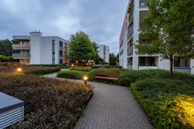 Modern bir yerleşim yerinin bahçesinde sakin bir akşam ortamı. Aydınlatılmış yollar ve aydınlatma huzur ortamını güçlendirir, karanlık çöktükten sonra açık havada karşılama alanı yaratır..