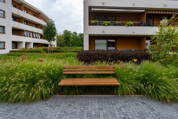 Süslü otların ve çalıların arasındaki ahşap bir bank sakinler için sakin bir rahatlama noktası sunuyor. Modern bir konut kompleksinin bahçe düzenlemesi, kentsel gelişimin güzel bir örneği.. 