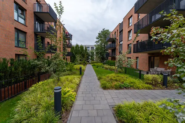 Çeşitli bitkiler ve balon ışıklarıyla çevrili asfalt bir yol, hoş yaya erişimi sağlar ve modern tuğla apartmanlar arasında doğrusal perspektif oluşturur.