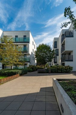 Güneşli bir günde, bir apartman kompleksi içinde sessiz bir avlu. Peyzaj alanı arasında kaldırımlı bir yürüyüş yolu, yeşil çimenler, ağaçlar ve yeni konut binaları arasında çalılıklar yer alıyor..