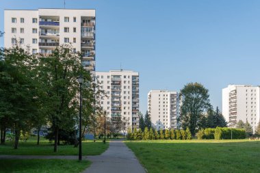 Yüksek yoğunluklu konutlarda yeşil çimenleri ve ağaçları olan büyük bir halk parkı. Yürüyen bir patika, arka planda uzun apartman blokları olan bu boş alanda esiyor..