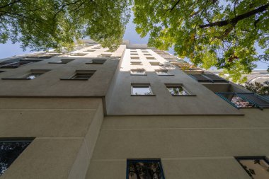 Düşük açılı bir perspektif yüksek bir konut binasının ön cephesine bakıyor. Manzara yerden gökyüzüne doğru uzanıyor, binanın tepesi yeşil ağaç yapraklarıyla çevrili..