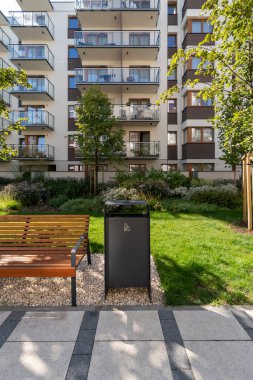 Konut kompleksli bir avluda kentsel konfor. Modern bir çöp tenekesi ve ahşap bir bank, toplum rahatlığı ve temizliği için peyzajlı bir kamusal alana yerleştirilir..