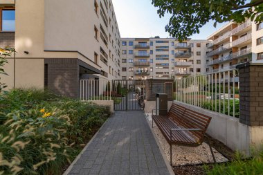 Güvenli kapısı olan modern bir yerleşim yerinin bahçesinde peyzaj yapılmış. Yaya geçidi ahşap bir bank ve yemyeşil bitkilerle çevrili bir çöp kutusuyla döşelidir..