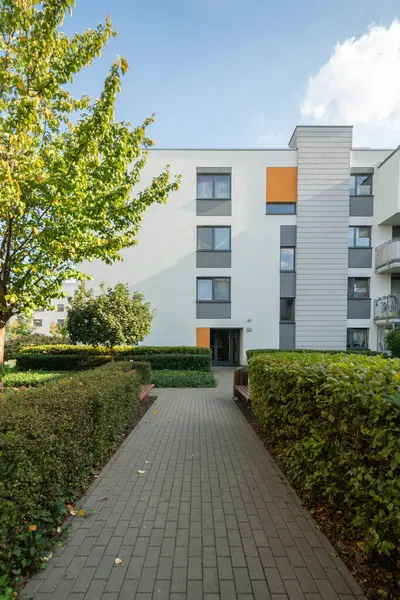 Yeni bir apartmanın mimari cephesi. Yeşil çitlerle ve park banklarıyla çevrili asfalt bir yürüyüş yolu, bu modern konut arazisinin girişine götürür..