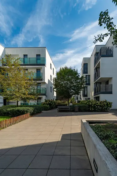Güneşli bir günde, bir apartman kompleksi içinde sessiz bir avlu. Peyzaj alanı arasında kaldırımlı bir yürüyüş yolu, yeşil çimenler, ağaçlar ve yeni konut binaları arasında çalılıklar yer alıyor..