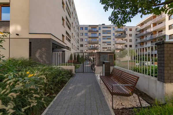 Güvenli kapısı olan modern bir yerleşim yerinin bahçesinde peyzaj yapılmış. Yaya geçidi ahşap bir bank ve yemyeşil bitkilerle çevrili bir çöp kutusuyla döşelidir..