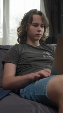 Genç bir adam evdeki kanepede rahatça otururken dizüstü bilgisayarında çalışır. Serbest çalışan, uzaktan çalışan ya da online kursa giden bir öğrenci olabilir..