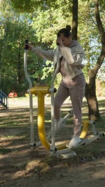 Spor giyimli bir kadın halka açık bir parkta eliptik bir makinede kardiyo çalışması yapıyor. Bu açık hava spor aletleri sağlıklı bir yaşam tarzı ve ücretsiz fitness eğitimi sağlıyor..
