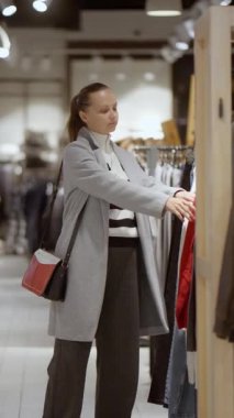 Bir kadın elbise raflarına bakar, modern bir perakende mağazasından alışveriş yapar. Geniş bir seçimden yeni giysiler seçme süreci modern moda ve tüketiciliği gösteriyor..