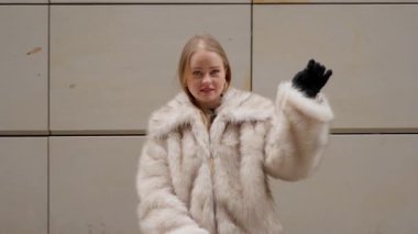 Şık, sahte kürklü, kendine güvenen sarışın bir kadın modern bir bina duvarına karşı poz veriyor. Döndü, kameraya baktı ve döndü. Şık kıyafetini sergiledi. Şehir ortamında zarifti..