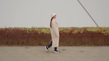 Sonbahar montu giymiş genç bir kadın modern bir bina boyunca yürür ve arkasına bakar. Şehir kaldırımında yürürken omzunun üzerinden bakan biri, düşünceli ya da meraklı bir an.