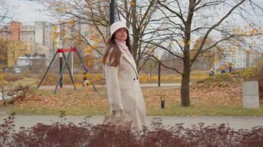 Şapkalı ve ceketli güzel bir kadın sonbahar sezonunda halka açık bir parkta rahatça dolaşır. Çocuk parkı ve konut binaları olan bir yolda yürüyen bir adam..