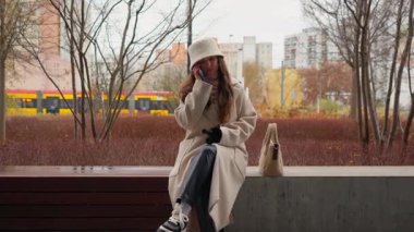 Sonbaharda parkta bir bankta otururken cep telefonuyla konuşan bir kadın. Arka planda tramvayla akıllı telefonuyla sohbet eden genç bir kız..