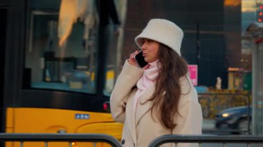 Akşamları toplu taşıma durağında bekleyen akıllı telefonuyla konuşan genç bir kadın. O kişisel konuşmasına odaklanırken arka planda bir tramvay geçiyor..