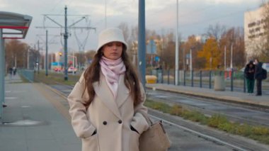 Paltolu ve şapkalı genç bir kadın soğuk bir sonbahar gününde tramvay durağında yürür ve nakil için bekler. İnsanlar, raylar ve mevsimlik şehir atmosferiyle kentsel sokak sahnesi.