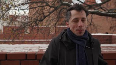 Bir adam utangaç bir gülümseme ile kameraya bakar, ağırlığını ayaktan ayağa kaydırır. Onun nazik ifadesi ve rahat eşarbı serin kış sokağına kişisel bir sıcaklık hissi veriyor..